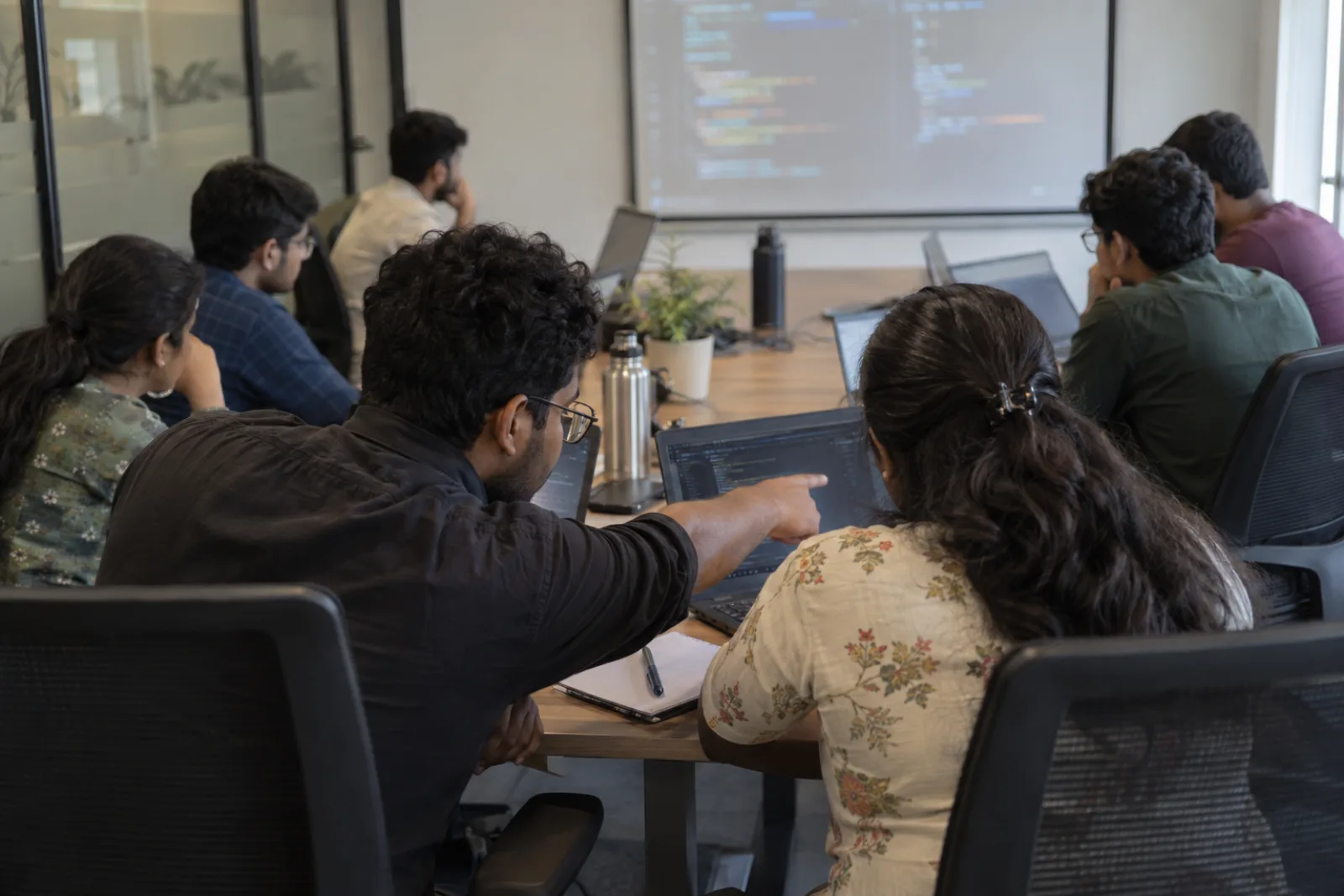 Engineers at laptops in a training room, one pointing at a colleague's screen, code on the projector (illustrative photo)