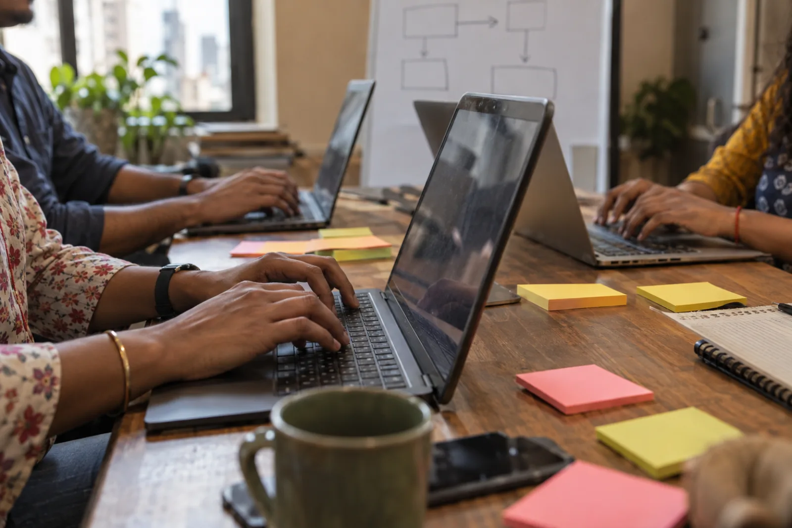 Hands typing on laptops around a wooden table scattered with sticky notes, a whiteboard flowchart behind (illustrative photo)